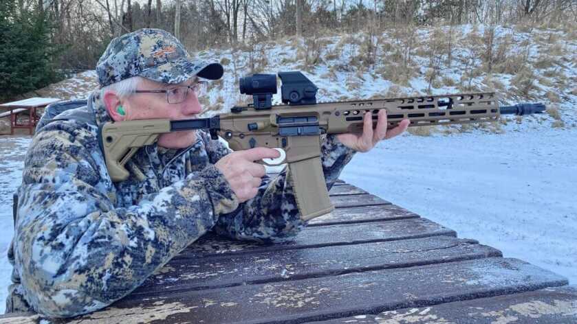 Man in camo jacket and hat shooting rifle mounted with Meproguard MMX3 Magnifier outside in the snow