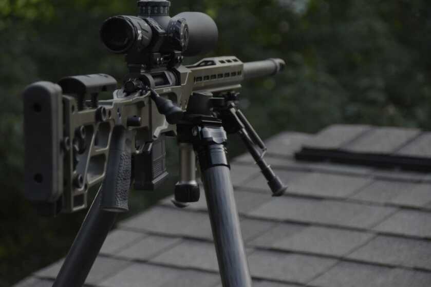 The rifle is mounted to a tripod and is perched on top of a shingled roof in an overwatch position in a moody environment.