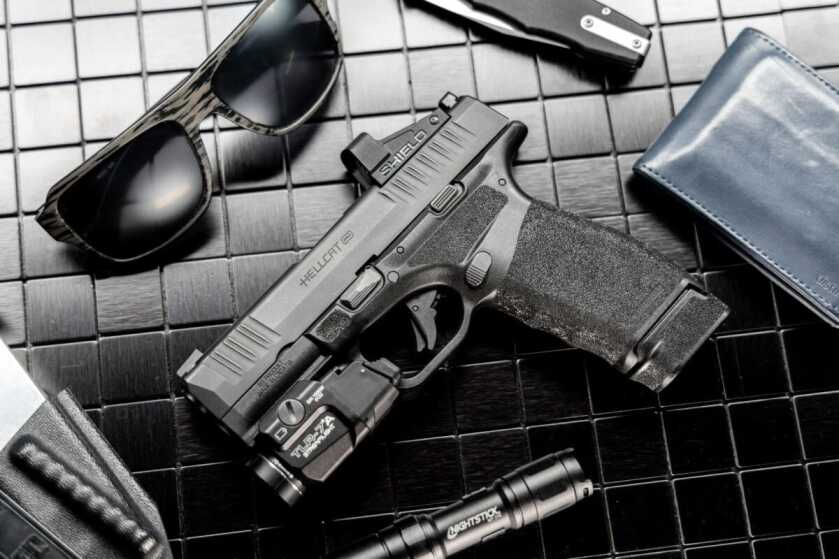 The Hellcat Pro lays on a nightstand surrounded by several other objects, including a pair of sunglasses, a wallet, and a pocket knife.