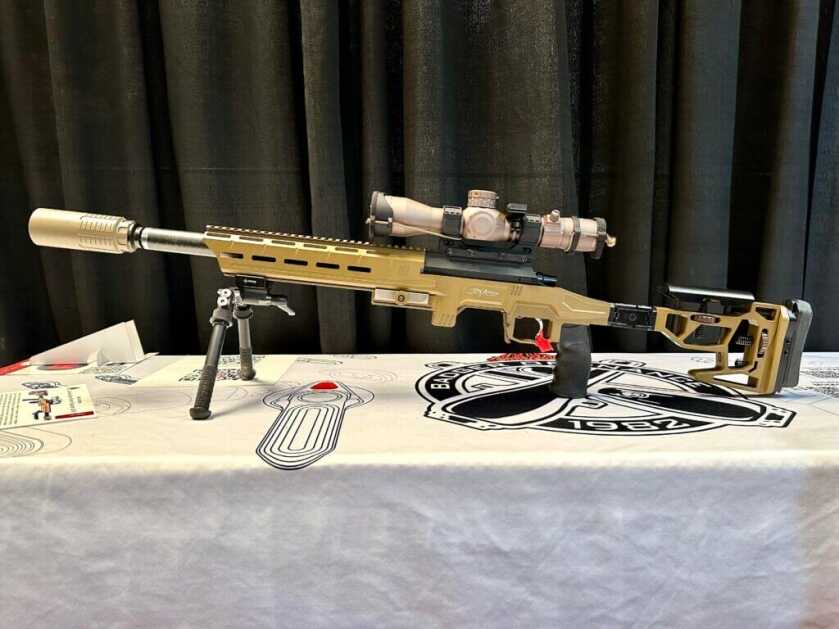 The fully assembled rifle sits on a bipod on a white table, facing to the left.