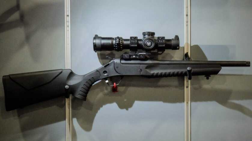 the LWC rifle is hanging on a wall facing to the right. It has a large optic mounted.