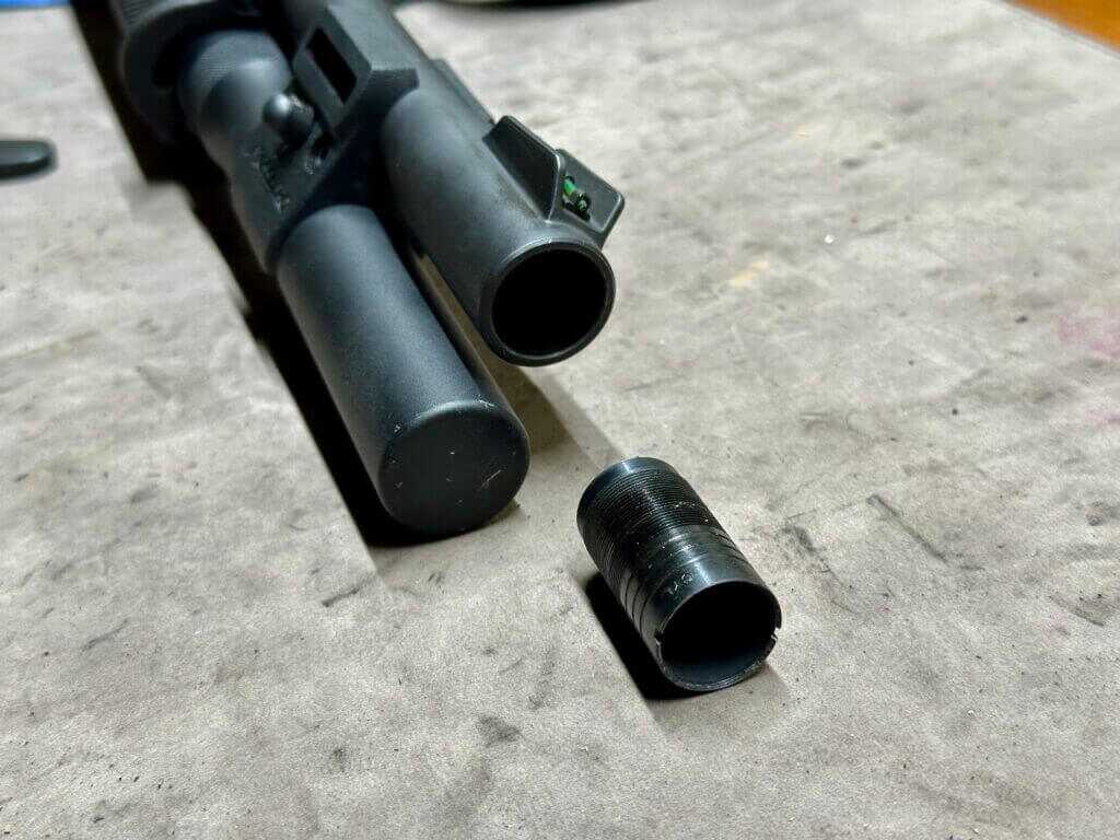 the choke tube is removed from a shotgun barrel and is being showcased on a tan mat, next to the gun.