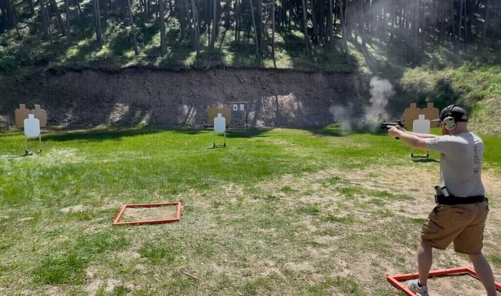 A man is shooting a gun at the gun range. An array of targets are in the background. MAX 