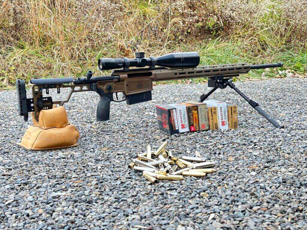 A rifle sits on a bipod and rear bag, behind an assortment of ammunition.