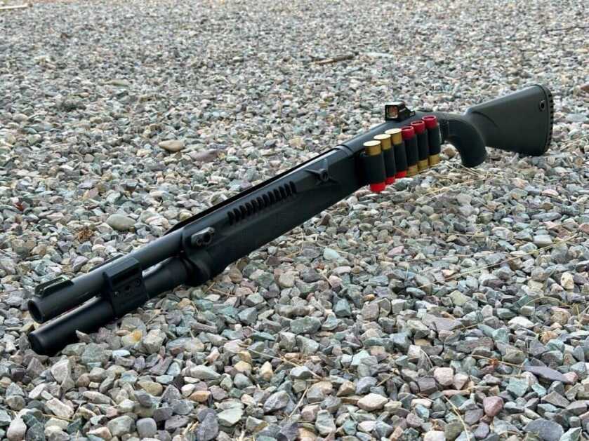 A shotgun lays in the gravel with shotgun shells strapped to its side.