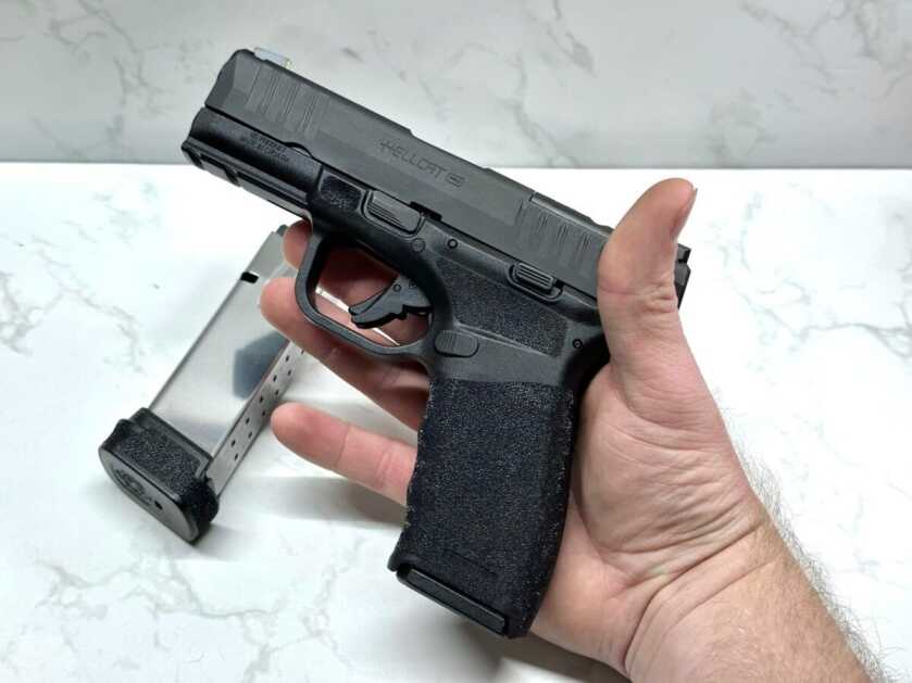 Holy Hellcat! Petite Pistol With Insane Upgraded Magazine Capacity A black handgun lays in the palm of the author's hand. The new Hellcat Pro extended magazine lays in the background against a white marble table top.