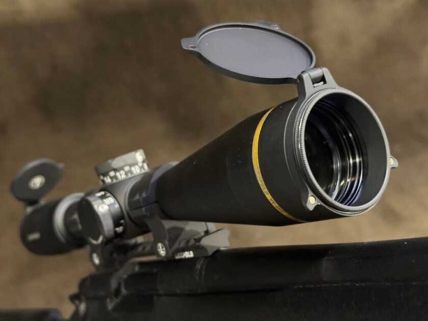 Peering at the rifle scope from the objective lense side, a scope cap is open and the shiny glass lens can be seen.