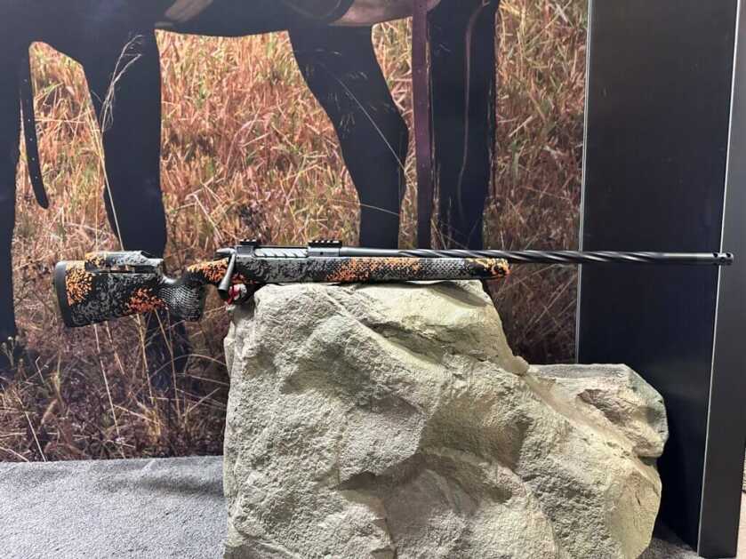 A rifle is balanced on a rock. It has a spiral fluted barrel and an orange camo pattern on the stock.