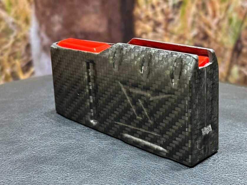A carbon fiber rifle magazine with a red follower sits on a black desk.