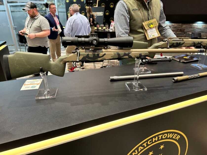 A rifle is staged on a tabletop, pointing to the right. People are in the background, walking around.