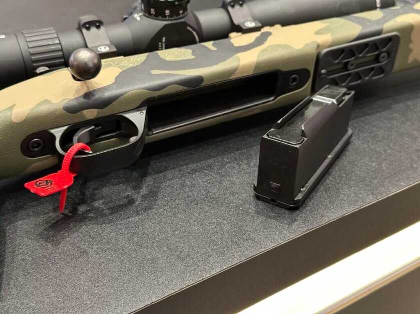 The bottom of a rifle is on display, showing a metal magazine, attachment points for tripods, and the profile of the rifle trigger.