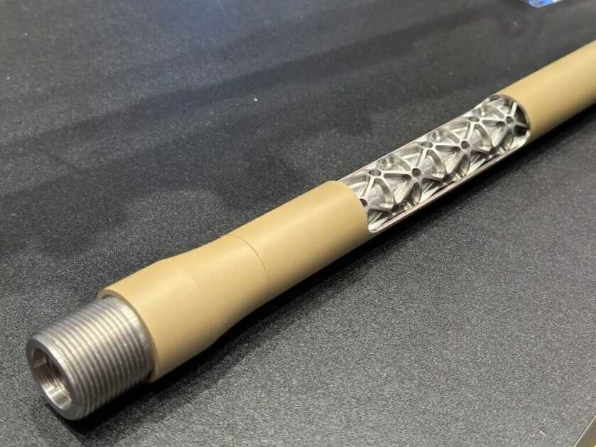 A rifle barrel lays on a black tabletop. There is a cutaway halfway down the barrel that shows intricate machinework under the outer sleeve.