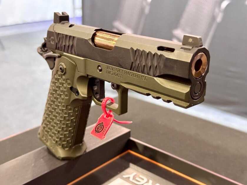 A green, black, and gold handgun is propped up on a stand. The gun faces toward us and to the right.