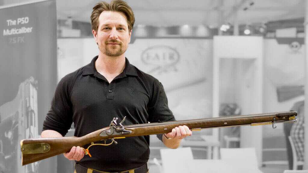 Photo of the upper portion of a man and he is holding the rifle in his hands with the rifle pointing to the right.