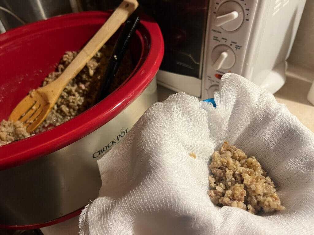 Bear cracklings draining through cheesecloth beside a red crockpot after rendering bear grease