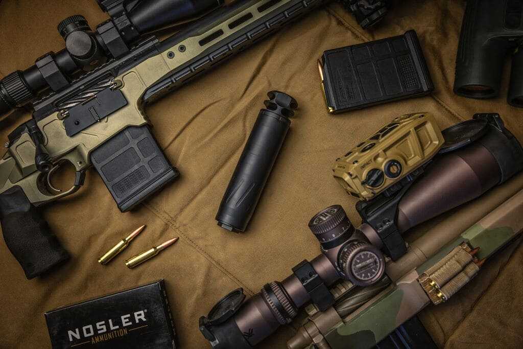 BANISH COMP 6.5 SS suppressor displayed with precision rifles, optics and 6.5mm ammunition.