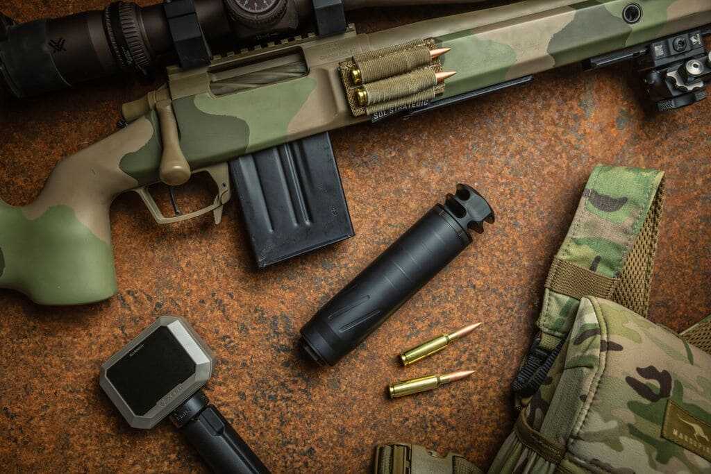BANISH COMP 6.5 TI suppressor displayed beside a scoped bolt-action rifle and hunting equipment.