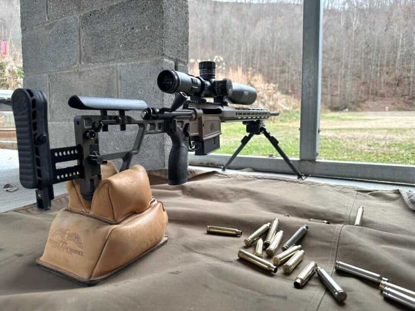 A rifle rests on bipod and rear bag, pointed down range at a gun range. 