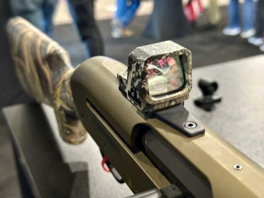 A red dot is mounted to a shotgun. Looking at the lense from the front, you can see the red reflective coatings.