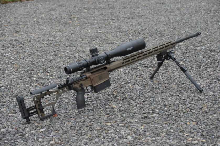 A tan rifle with a large optic is posed in the gravel.