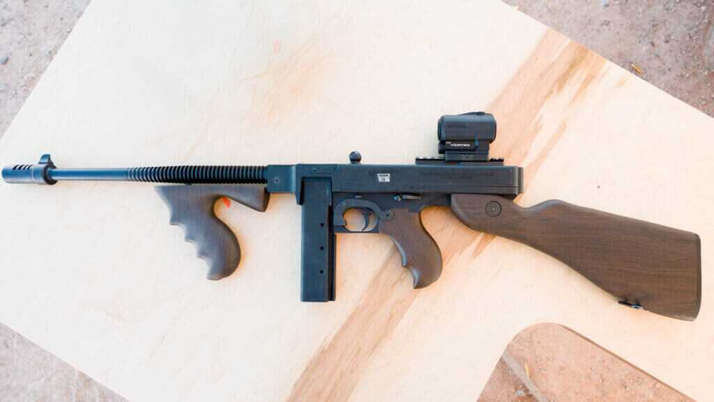 The whole tommy gun on a table pointing to the left, left side up.