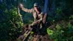 The author in the woods at near dark with a dead bear at his feet holding a cell phone flashlight up to the side for illumination on himself and the bear.