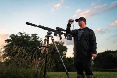 Hunter using the Athlon Cronus ATS P-MINI 250S thermal monocular while observing a field beside a suppressed rifle mounted on a tripod.
