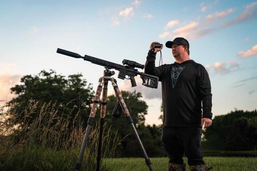 Hunter using the Athlon Cronus ATS P-MINI 250S thermal monocular while observing a field beside a suppressed rifle mounted on a tripod.