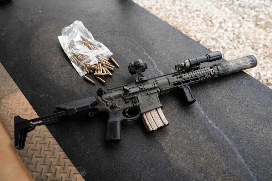Primary Weapons Systems MK107 MOD 2-M .300 Blackout PDW with a suppressor, folding stock, red dot optic and loaded magazine resting on a shooting bench beside ammunition.