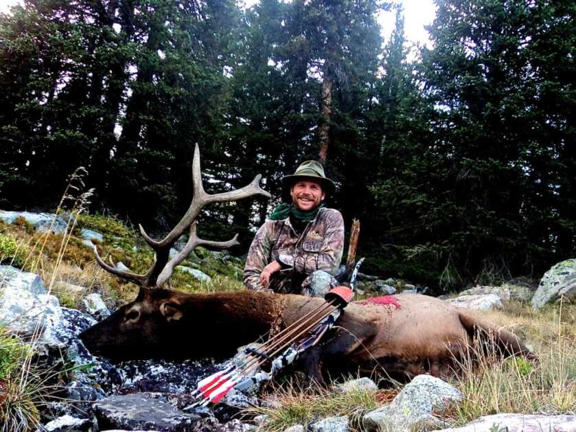 TAKE THE SHOT: A traditional bowhunter is offered a mid-range shot at a moving bull elk. Should he take the shot? Hunter posing with his elk, longbow, and quiver in a forest clearing