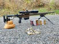 A rifle sits on a bipod and rear bag, behind an assortment of ammunition.