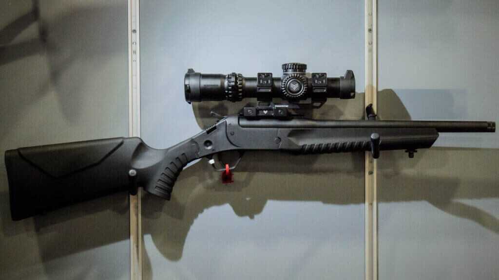 the LWC rifle is hanging on a wall facing to the right. It has a large optic mounted.