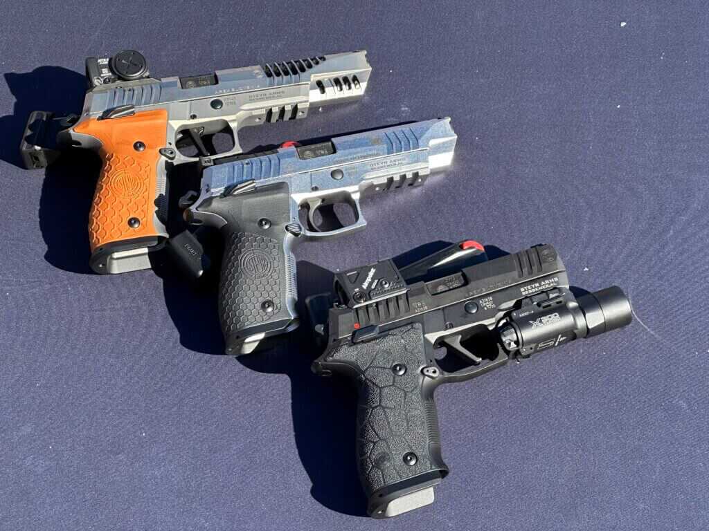 There are three pistols on a blue tabletop. the top one is silver with orange grips, the middle one is silver with black accents, and the bottom one is black.