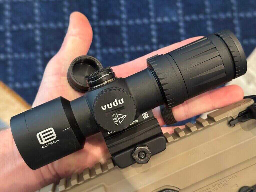 A compact optic lays in the palm of a man's hand and the elevation turret is uncapped.