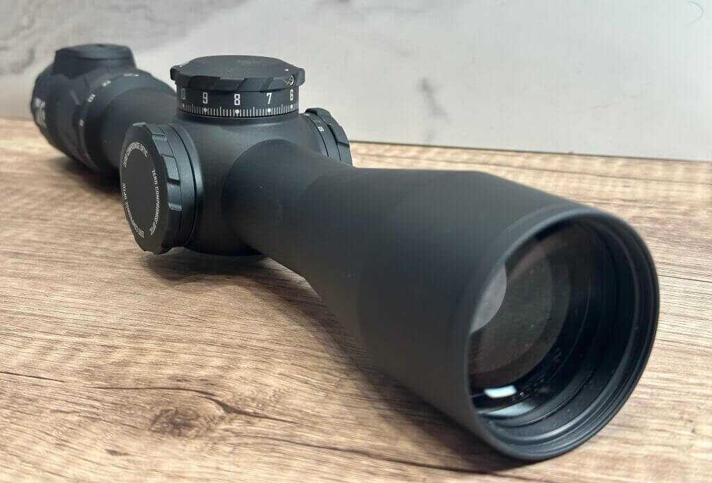 View of a rifle scope pointing toward us. We see the objective lense and sxope body behind it.