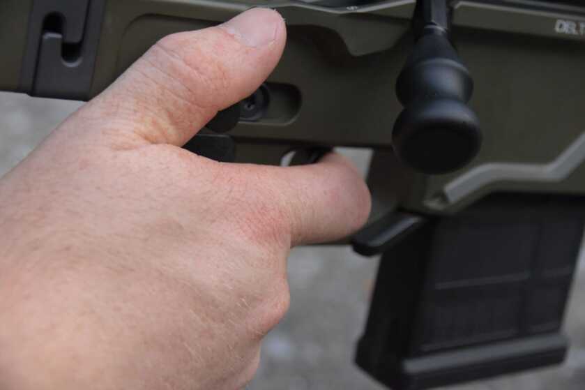 A hand is placed on the rifle's grip, finger on the trigger and thumb on the built in thumb-stop. 