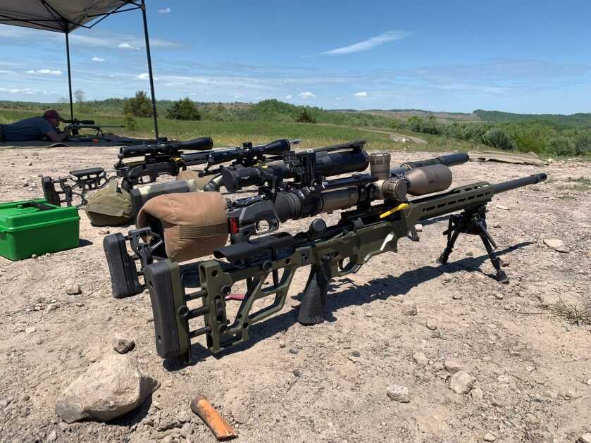 Several rifles are stood in a line on bipods at a shooting event while a man is prone in the background, shooting his gun.