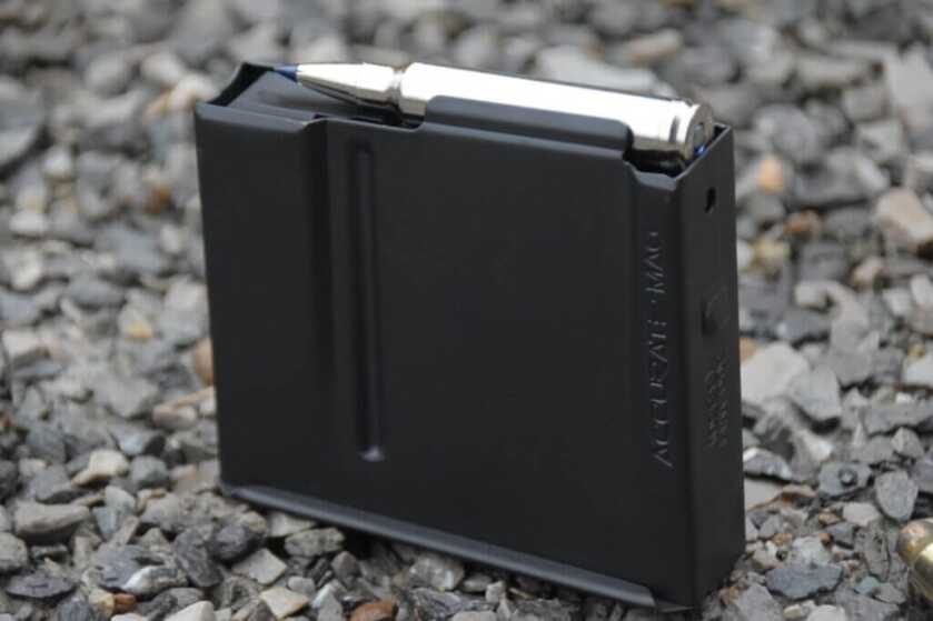 A metal rifle magazine with one cartridge in it sits on gravel.