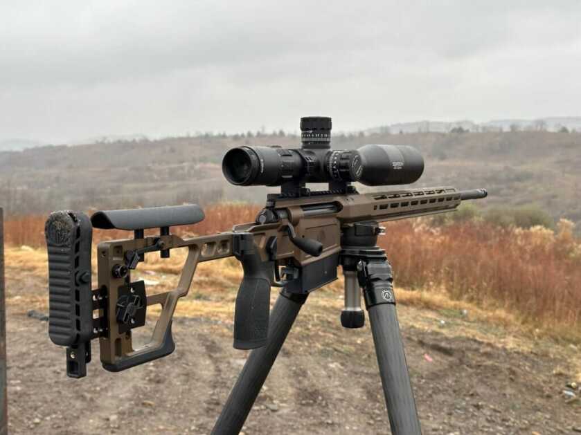 A brown rifle is positioned on a tripod for standing positional shooting.