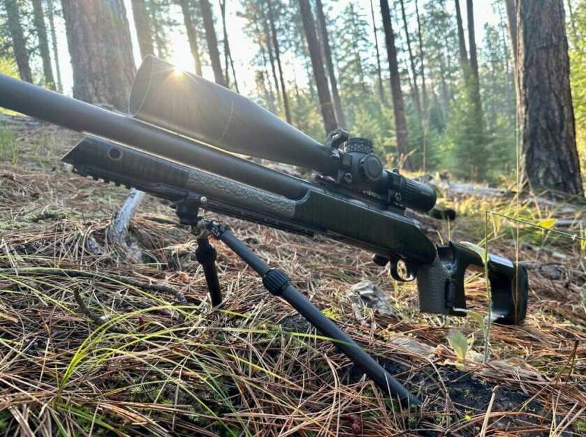 A black rifle is posed on a green, treed hillside.