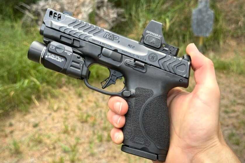 Hand holding Smith & Wesson M2.0 Carry Comp pistol with Defender-ST red dot and Streamlight weapon-mounted-light outside near target and grass