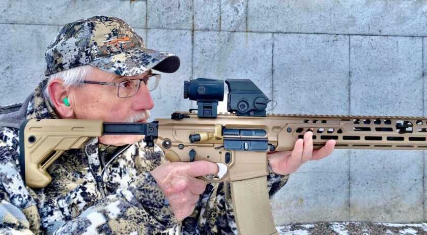 Man in camo shooting Sig MCX-LT rifle with Meprolight MMX3 Magnifier and Tru-Vision red dot in front of cement wall