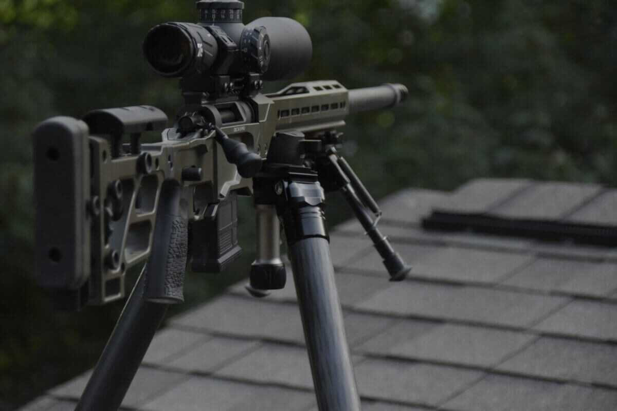 The rifle is mounted to a tripod and is perched on top of a shingled roof in an overwatch position in a moody environment.