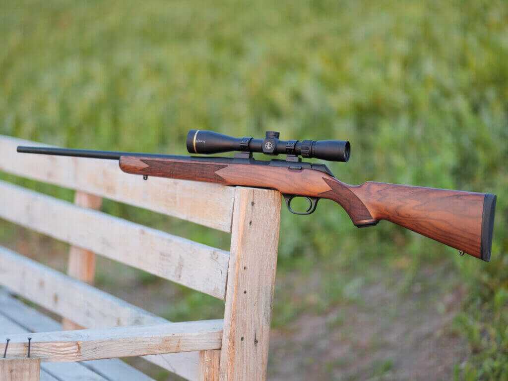 profile of the entire rifle of the gun's left side with the barrel pointing to the left. The rifle has a wood stock, black hardware, and scope.