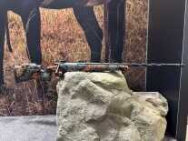 A rifle is balanced on a rock. It has a spiral fluted barrel and an orange camo pattern on the stock.
