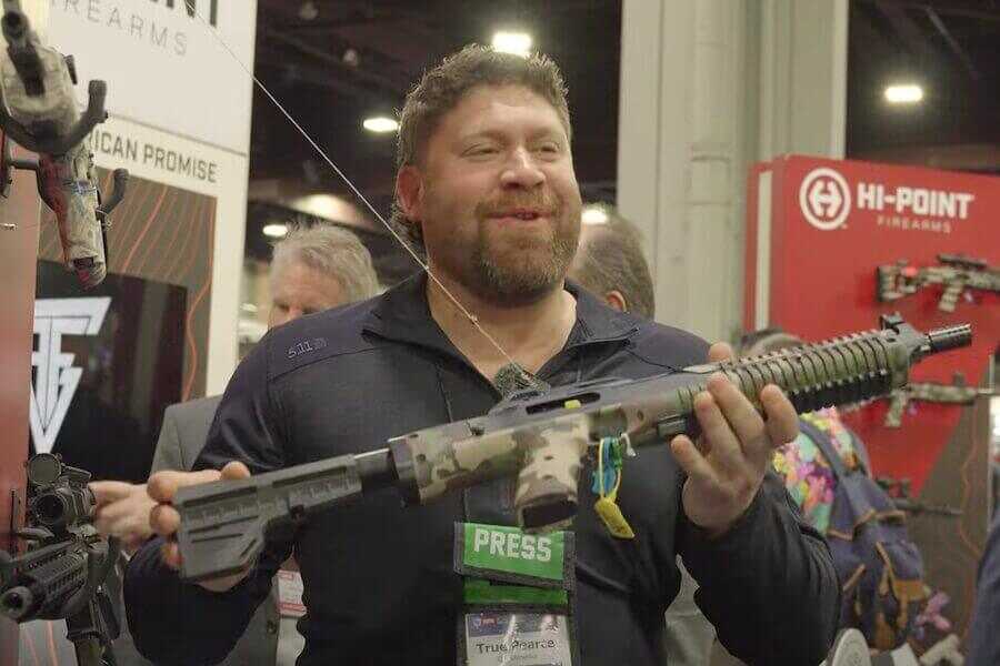 True holding up a Woodland camo Hi-Point 995P.