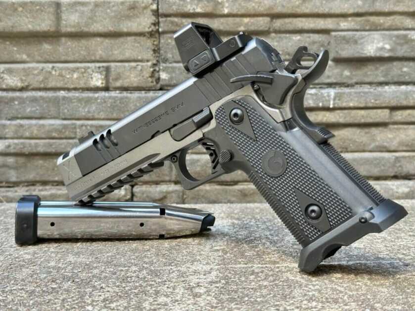 A black and silver handgun with an optic is staged by a silver magazine in front of a stone backdrop. Witness2311 Match X