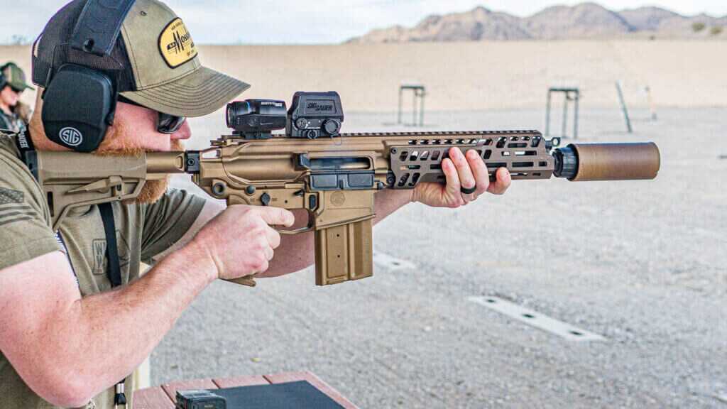 A closeup view of a man holding the M7 rifle, ready to fire. The background is a gravel shooting range with mountains in the background.