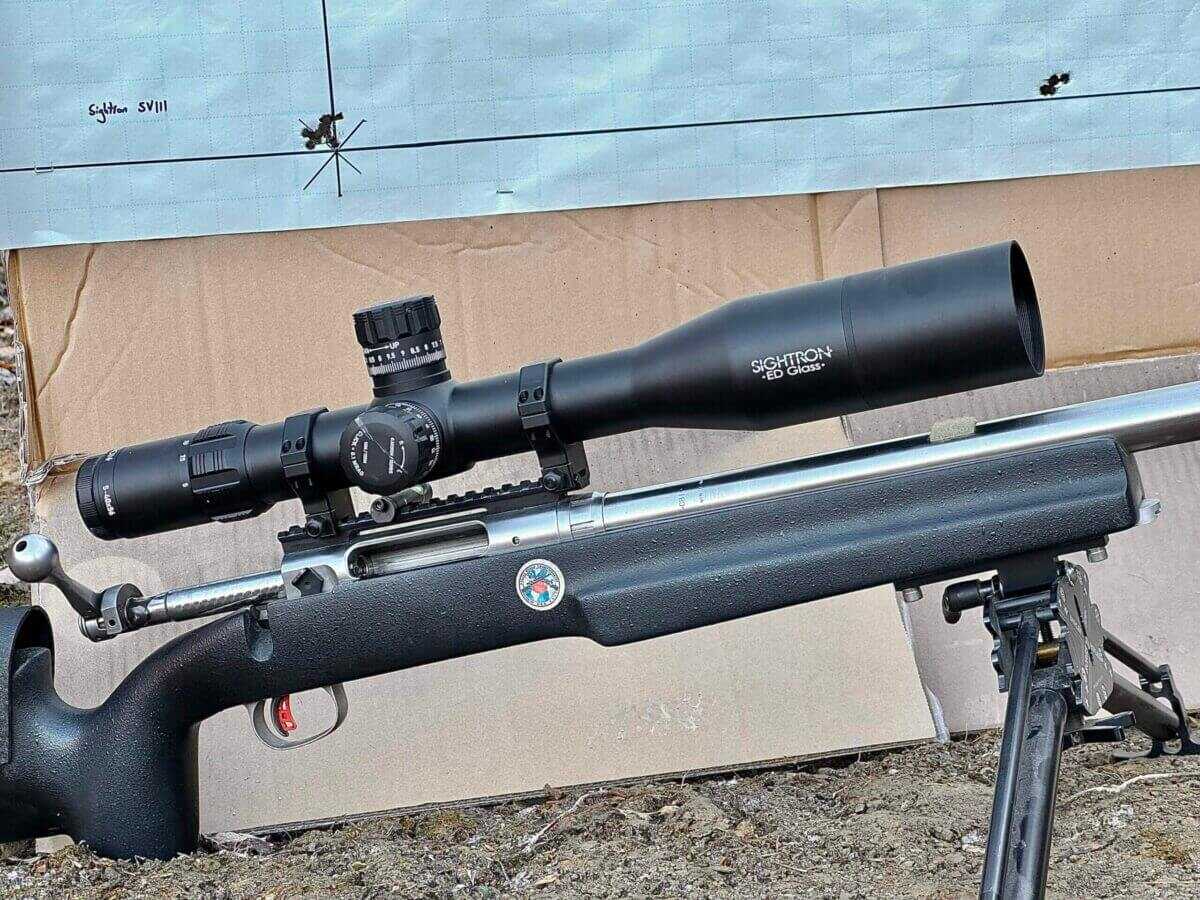 Sightron SVIII A target with bullet holes in it sits behind a target rifle with a large optic on it.