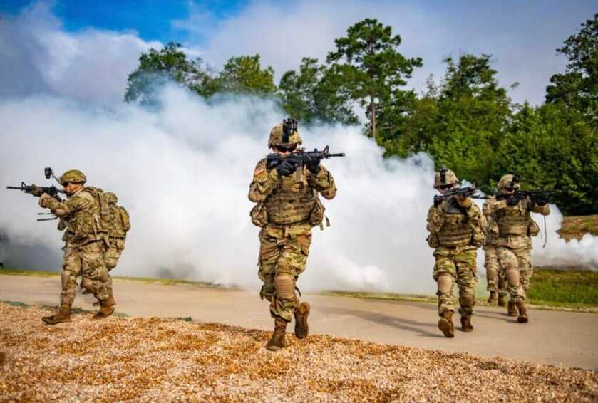 4 US Army soldiers in full gear with rifles moving in front of smoke and trees